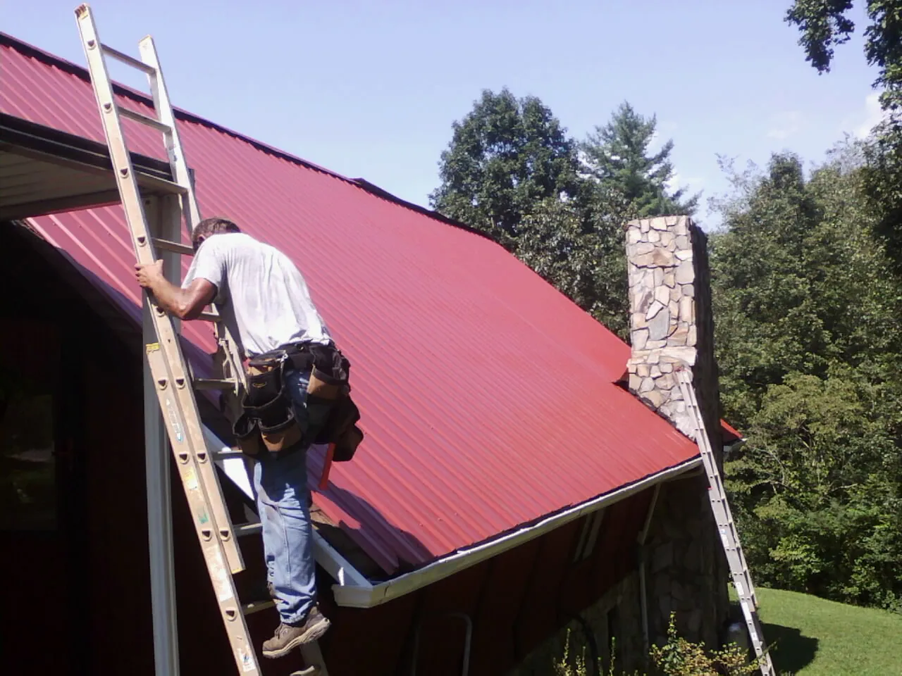 Roofer on a ladder beside a red metal roof and stone chimney — Soffit & Fascia Repair by Roofing Brothers in Libertyville